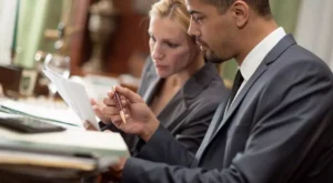 Imagem de homem negro e mulher branca de ternos lendo documento de publicação no Diário Oficial.