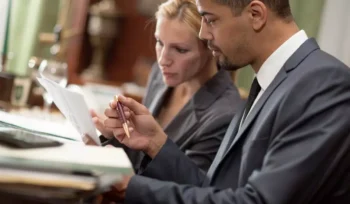 Imagem de homem negro e mulher branca de ternos lendo documento de publicação no Diário Oficial.