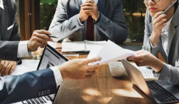Imagem de pessoas de terno em volta de uma mesa, com uma mulher entregando papel a um homem, explicando o que é pregão.