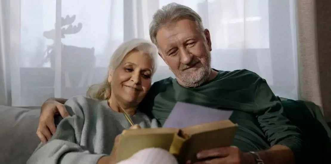 Imagem de um casal de idosos brancos sentados no sofá com livro na mão após aposentadoria proporcional.
