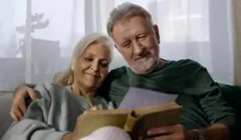 Imagem de um casal de idosos brancos sentados no sofá com livro na mão após aposentadoria proporcional.