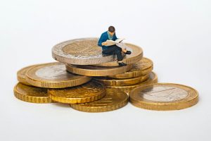 Várias moedas em um fundo branco com um boneco de um homem sentado em cima delas, lendo um papel e simulando um ativo realizável em longo prazo