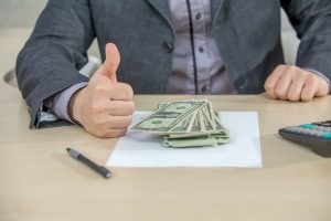 Pessoa fazendo sinal de positivo ao lado de notas de dólar, representando a repatriação de recursos.
