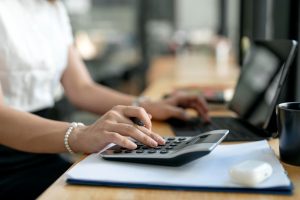 Mulher sentada em frente ao computador, com uma mão no teclado e outra na calculadora ao lado, calculando o balanço patrimonial e DRE