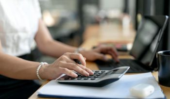Mulher sentada em frente ao computador, com uma mão no teclado e outra na calculadora ao lado, calculando o balanço patrimonial e DRE