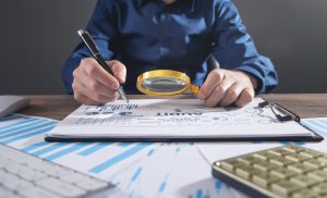 Pessoa de camisa social azul segurando uma lupa e uma caneta analisando documentos em uma auditoria financeira.